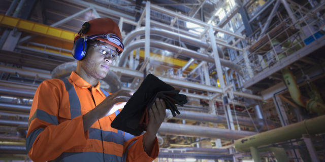 Worker in safety gear using a tablet in an industrial plant with pipes and machinery.
